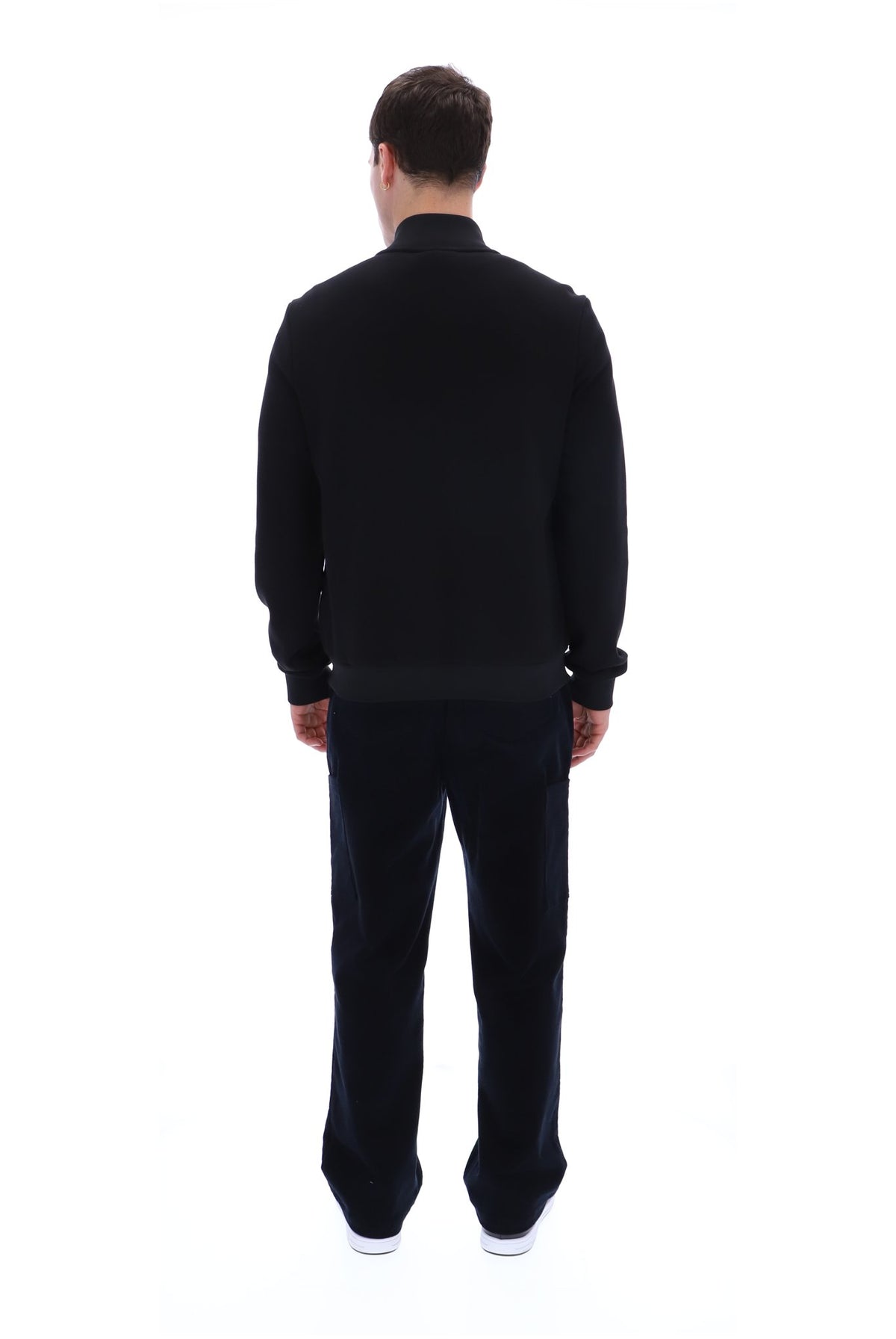 A man viewed from behind wearing a black long-sleeve knit top and matching black trousers, standing against a plain white studio background.