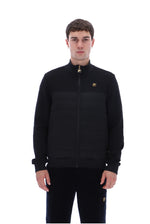 A man wearing a black zip-up jacket with horizontal quilting on the lower half and a small gold logo on the left chest, paired with black pants, against a plain white background.