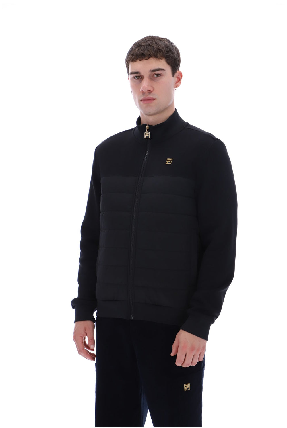A man wearing a black full-zip puffer jacket with a stand-up collar and a small gold logo on the chest, paired with black pants, photographed against a plain white background.