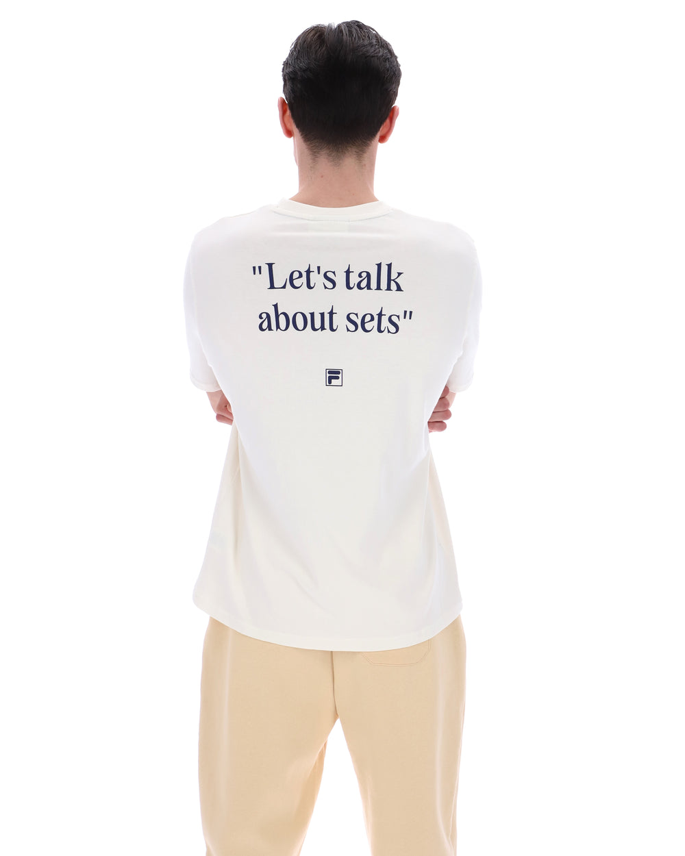 Person wearing a white t-shirt with text reading "Let's talk about sets" on the back, with a small black FILA logo standing against a white background