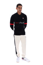 A man modeling a black long-sleeve polo shirt with red and white accents, paired with off-white track pants and white sneakers, against a plain white background.