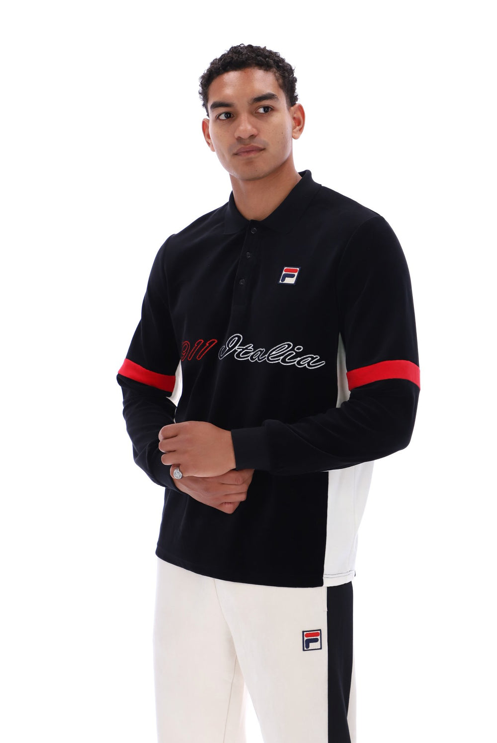 A man modeling a black Fila long-sleeve polo shirt with red and white accents, paired with white pants, against a plain white background.