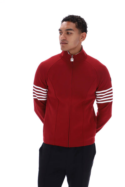 A man wearing a red zip-up track jacket with white stripes on the sleeves, paired with black pants, photographed against a solid white background.
