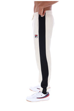 Side view of a person wearing off-white sweatpants with a bold black vertical stripe down the side and a small Fila logo patch near the upper thigh, paired with white sneakers on a plain white background.