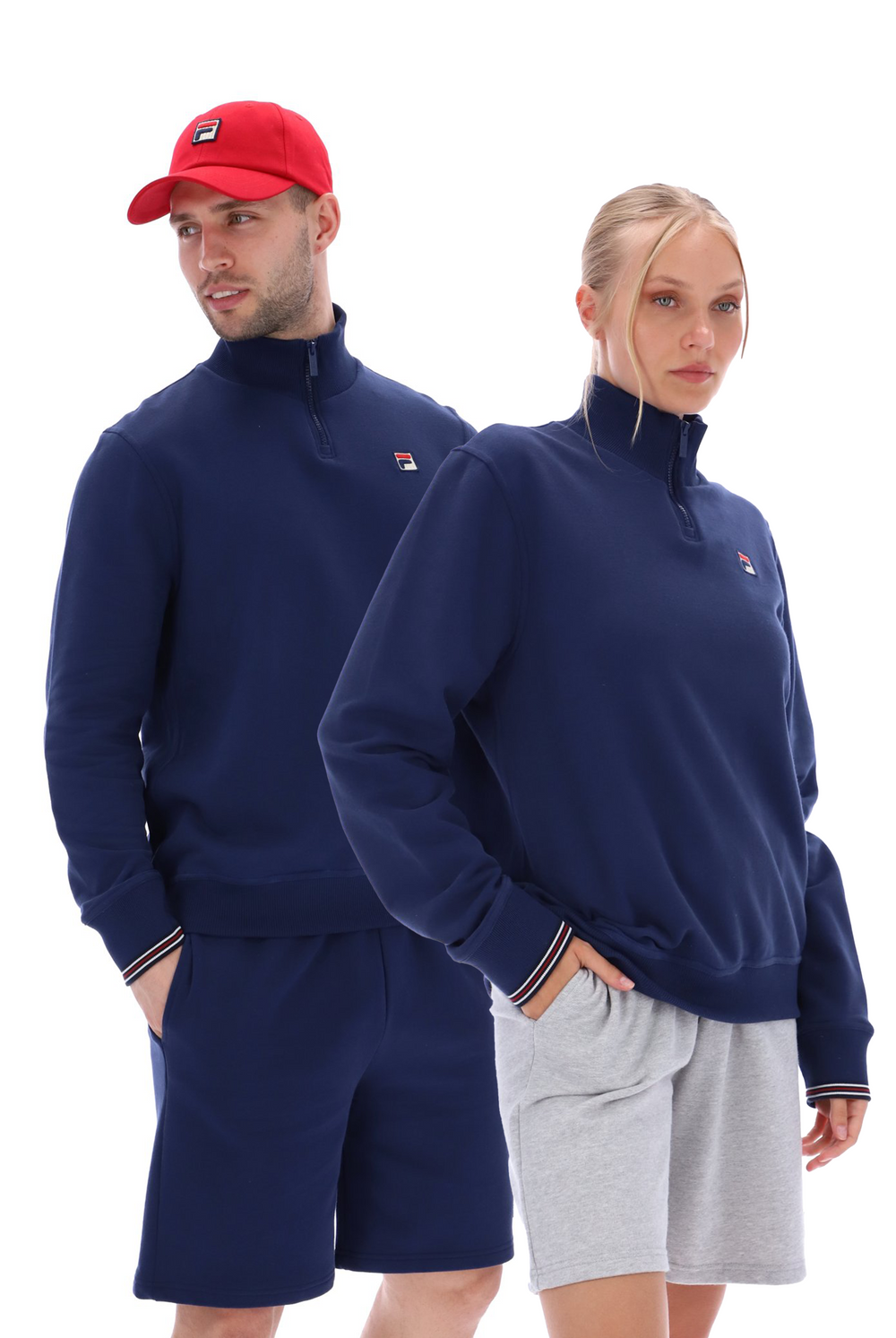 A male model wearing a red cap and a blue quarter zip sweatshirt with white stripes on the cuffs, and a female model wearing a blue quarter zip sweatshirt with white stripes on the cuffs and gray shorts.