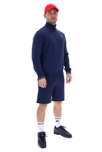 Man wearing a navy quarter zip jumper with a small FILA logo on the chest, paired with grey FILA jogger shorts, against a white background