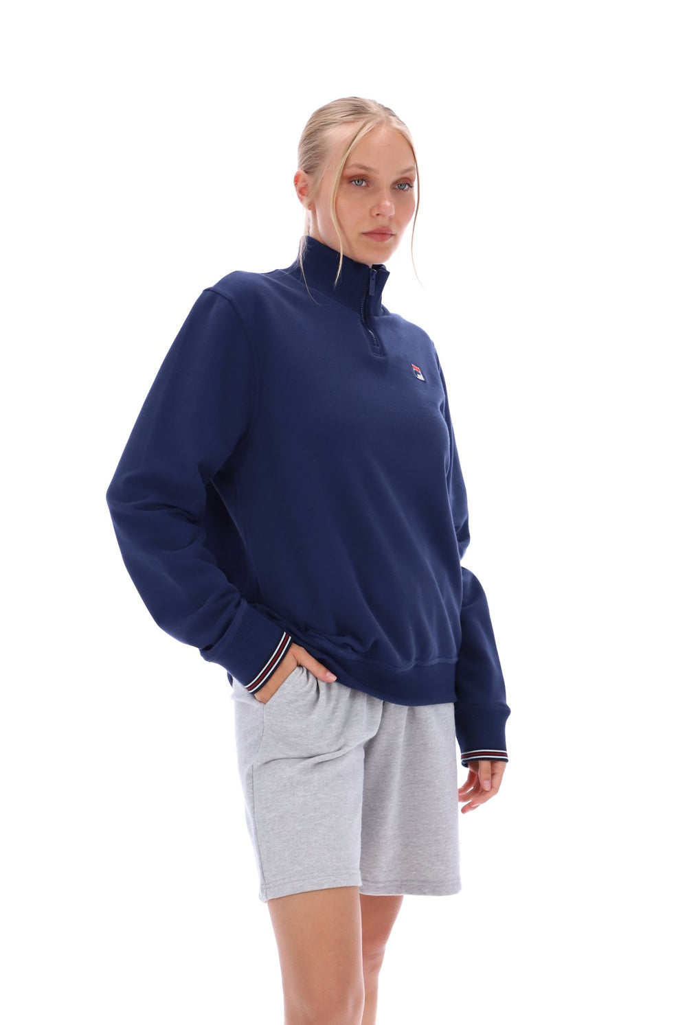 Woman wearing a navy quarter zip jumper with a small FILA logo on the chest, paired with grey FILA jogger shorts, against a white background