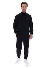 A man modeling a black quarter-zip fleece jacket and matching black jogger pants, featuring a small red, white, and blue Fila logo on the left chest, paired with white sneakers, against a plain white background.