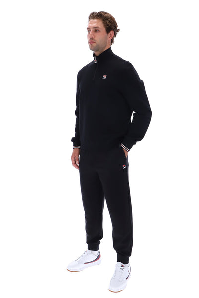 A man modeling a black Fila quarter-zip sweatshirt and matching black sweatpants with white sneakers, shown against a plain white background.