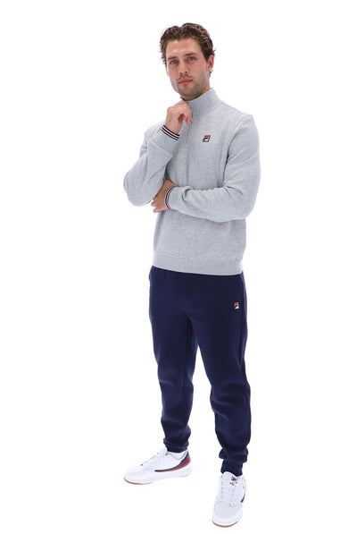 A man modeling a light gray heathered quarter-zip sweatshirt and navy blue joggers with white sneakers, featuring the Fila logo on the chest and thigh.