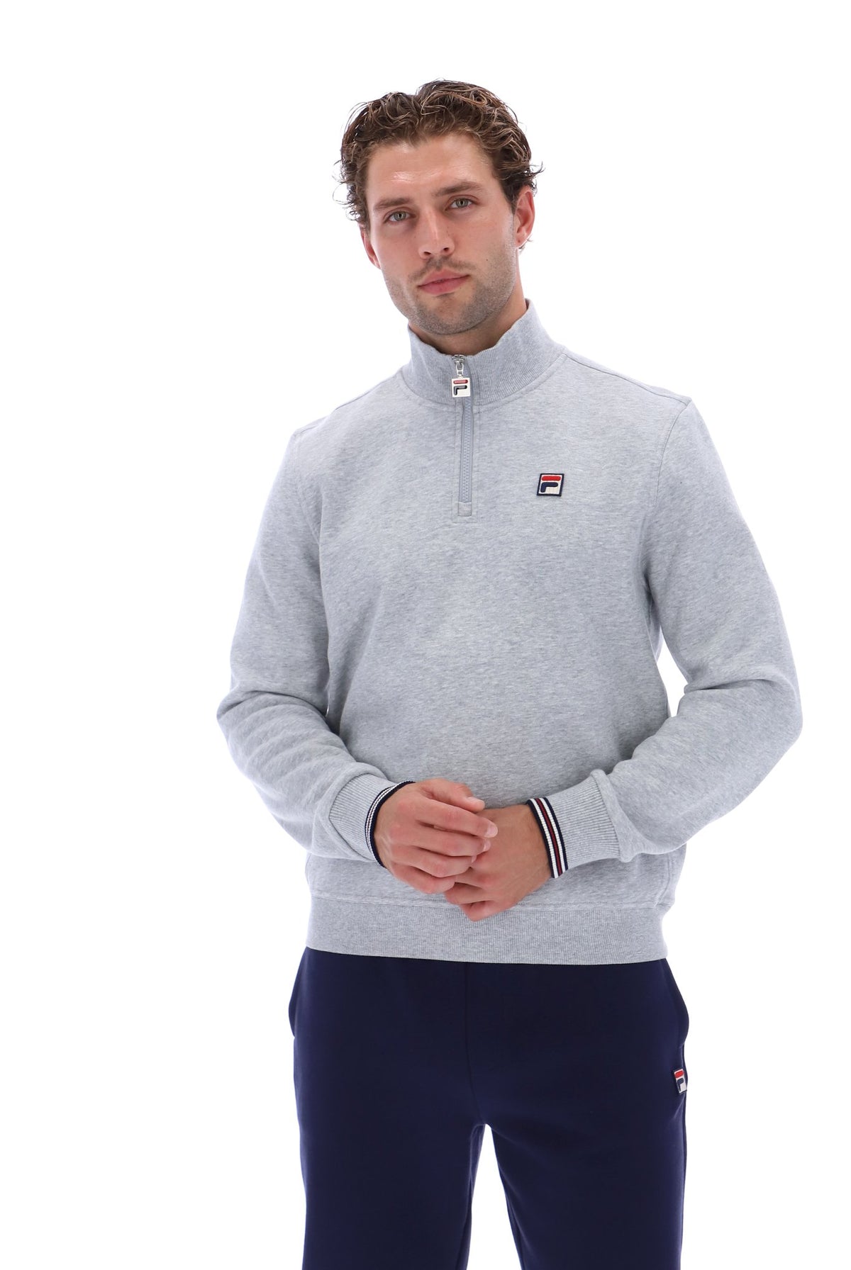 A man wearing a light gray heathered quarter-zip sweatshirt with a small Fila logo on the chest, paired with navy pants, against a plain white background.