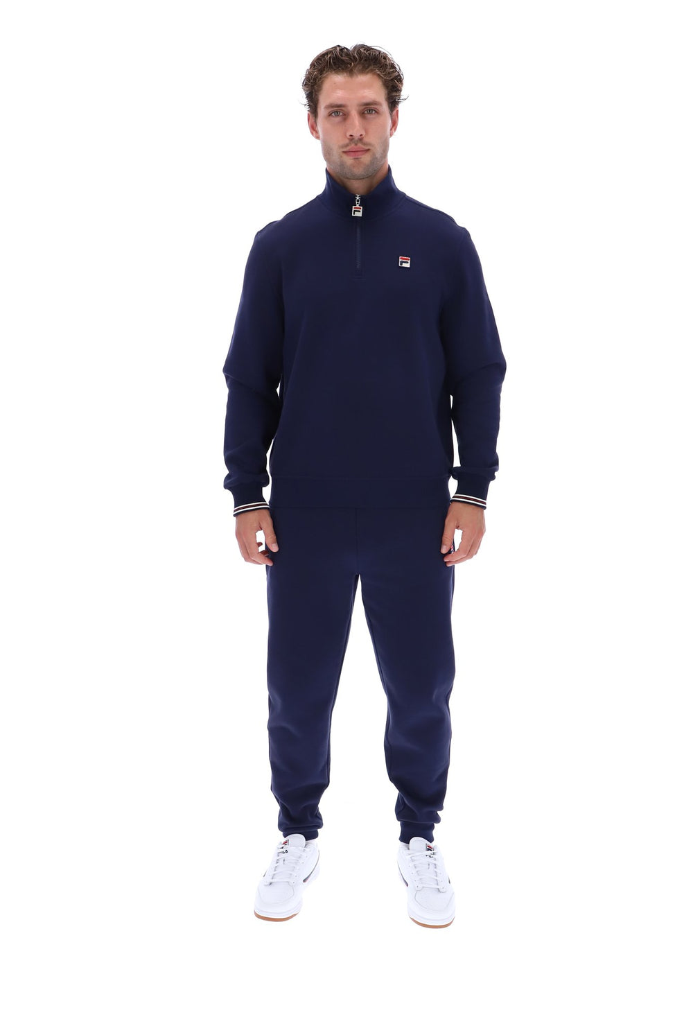 A man modeling a navy blue quarter-zip sweatshirt and matching sweatpants set, featuring a small red and white Fila logo on the chest, against a plain white background.