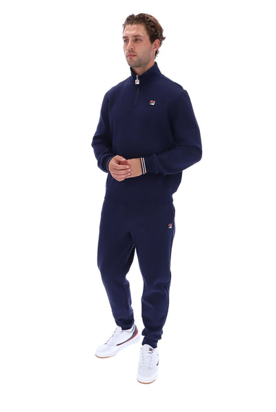 A man modeling a navy blue tracksuit with a quarter-zip pullover and matching joggers, featuring small red and white Filson logos on the chest and thigh, paired with white sneakers, against a plain white background.