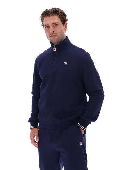A man wearing a navy blue Fila quarter-zip sweatshirt and matching sweatpants, standing against a solid white background.