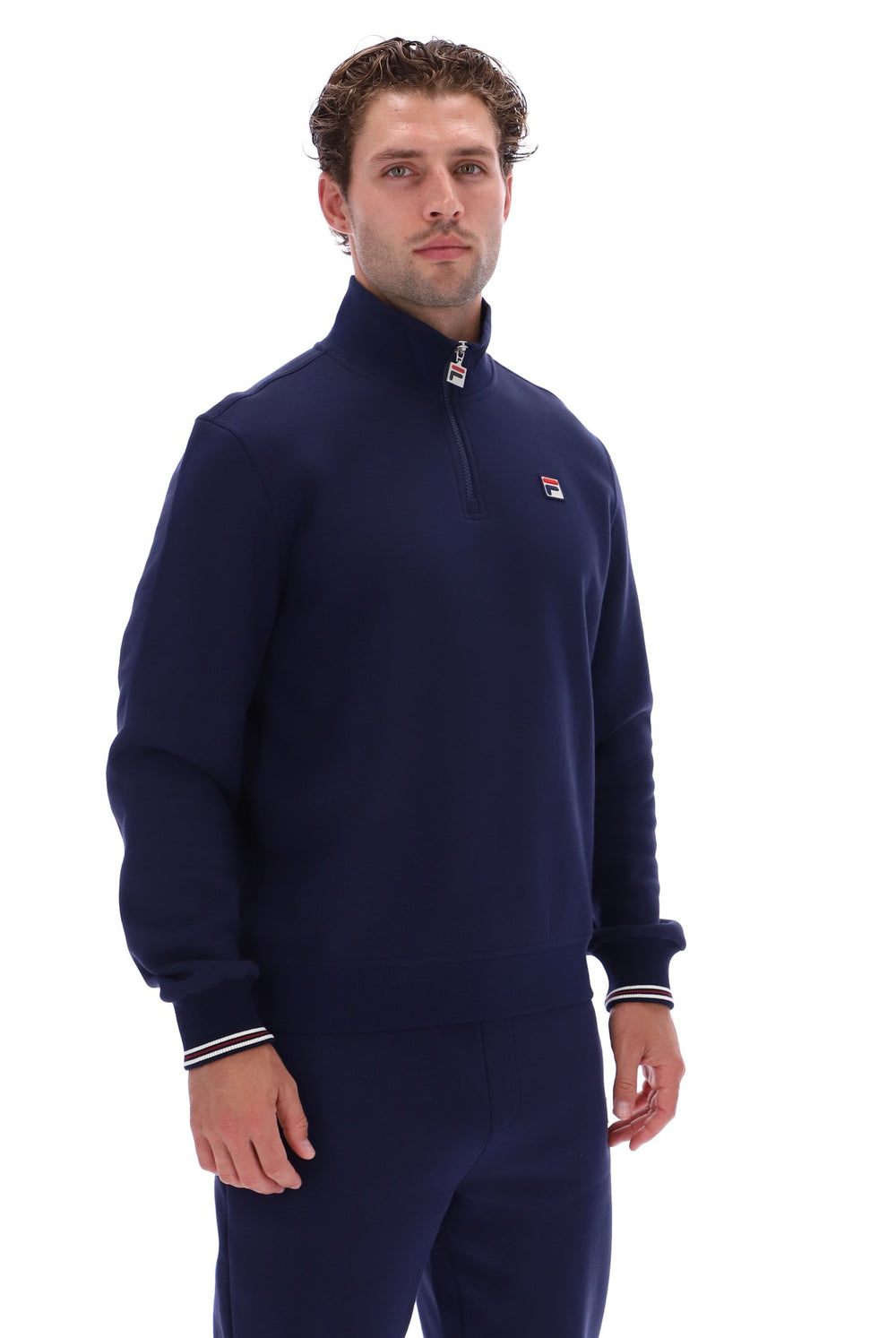 A man wearing a navy blue quarter-zip sweatshirt and matching sweatpants with white and red striped cuffs, featuring a small Fila logo on the chest.