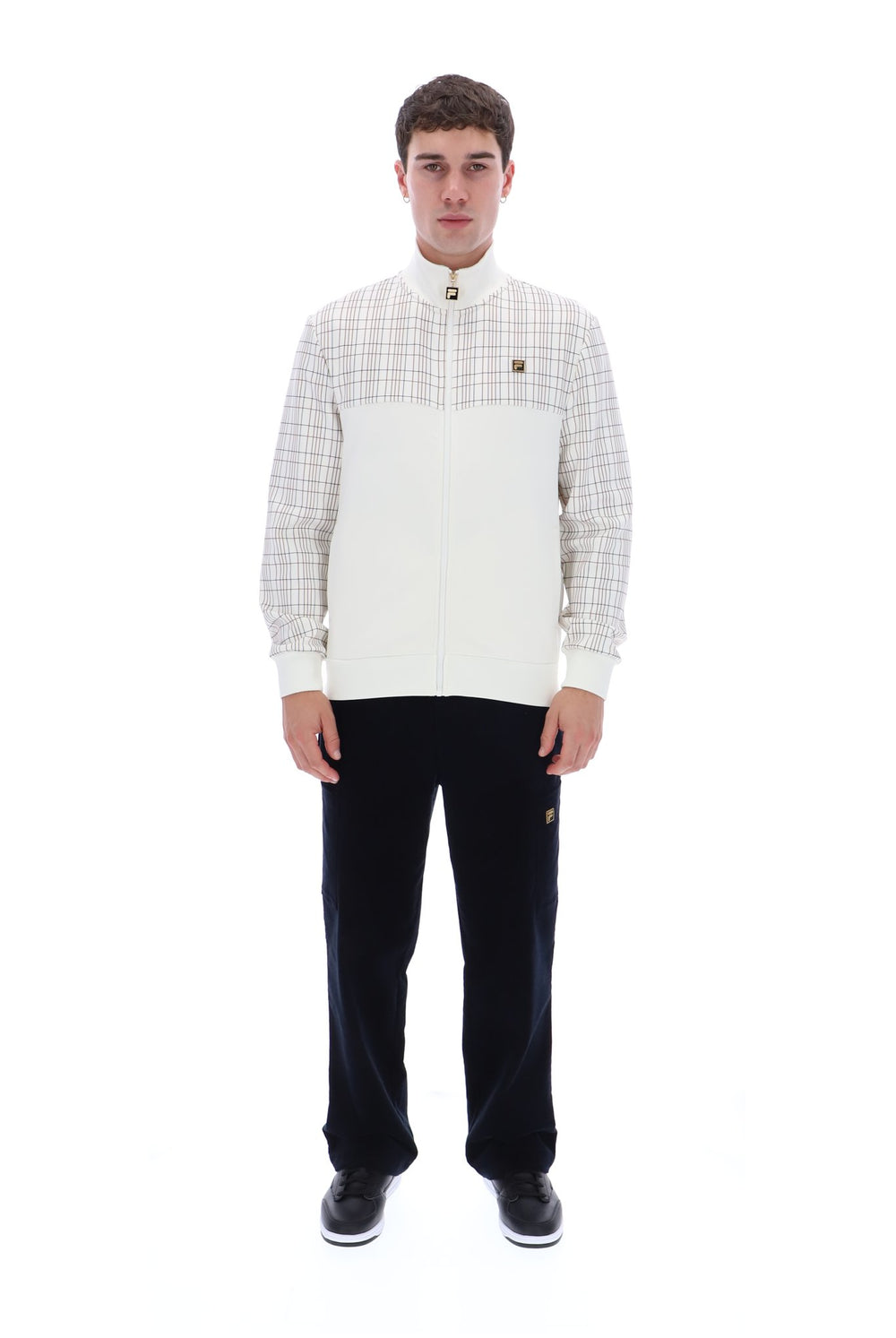 A man modeling a white and beige plaid zip-up jacket with a high collar, paired with black trousers and black sneakers, against a solid white background.