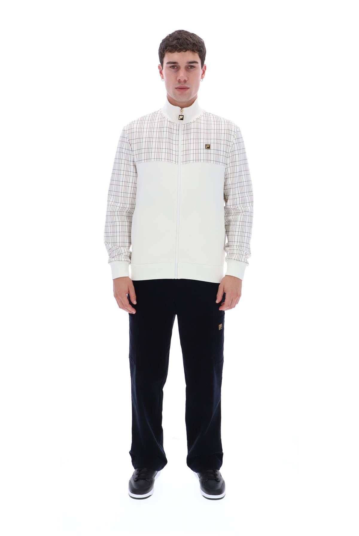 A man modeling a white and beige plaid zip-up jacket with a high collar, paired with black trousers and black sneakers, against a solid white background.