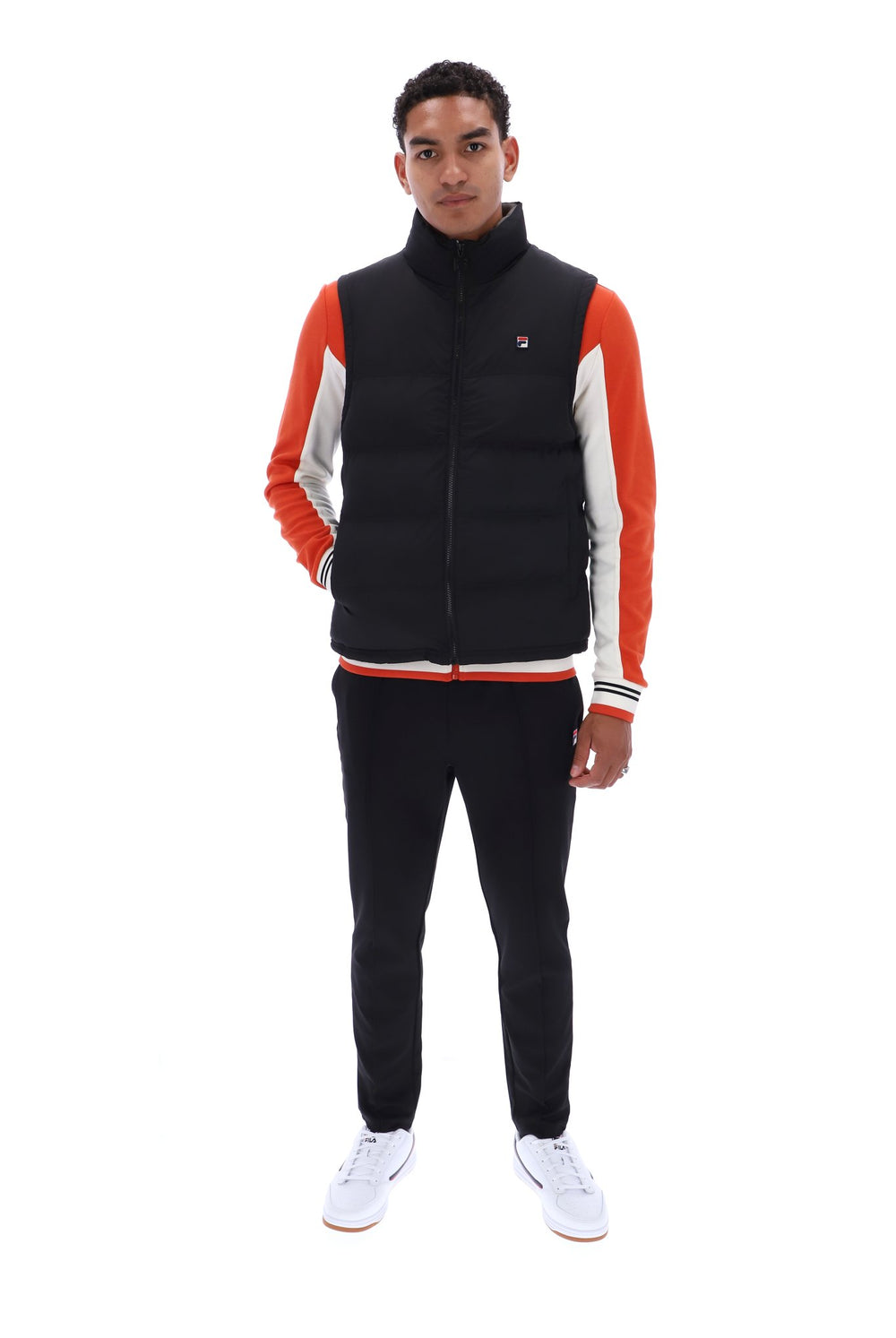 A man modeling a black puffer vest with orange and white color-blocked sleeves, paired with black sweatpants and white sneakers, against a solid white background.