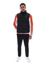 A man modeling a black puffer vest with orange and white color-blocked sleeves, paired with black sweatpants and white sneakers, against a solid white background.