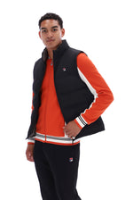 A man modeling a black and orange Fila tracksuit with a black quilted vest, orange zip-up jacket, and black sweatpants against a white background.
