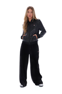 A woman modeling a black Fila tracksuit, featuring a faux-leather zip-up jacket with a small red and white logo on the chest, paired with wide-leg black velour sweatpants, shot against a solid white background.