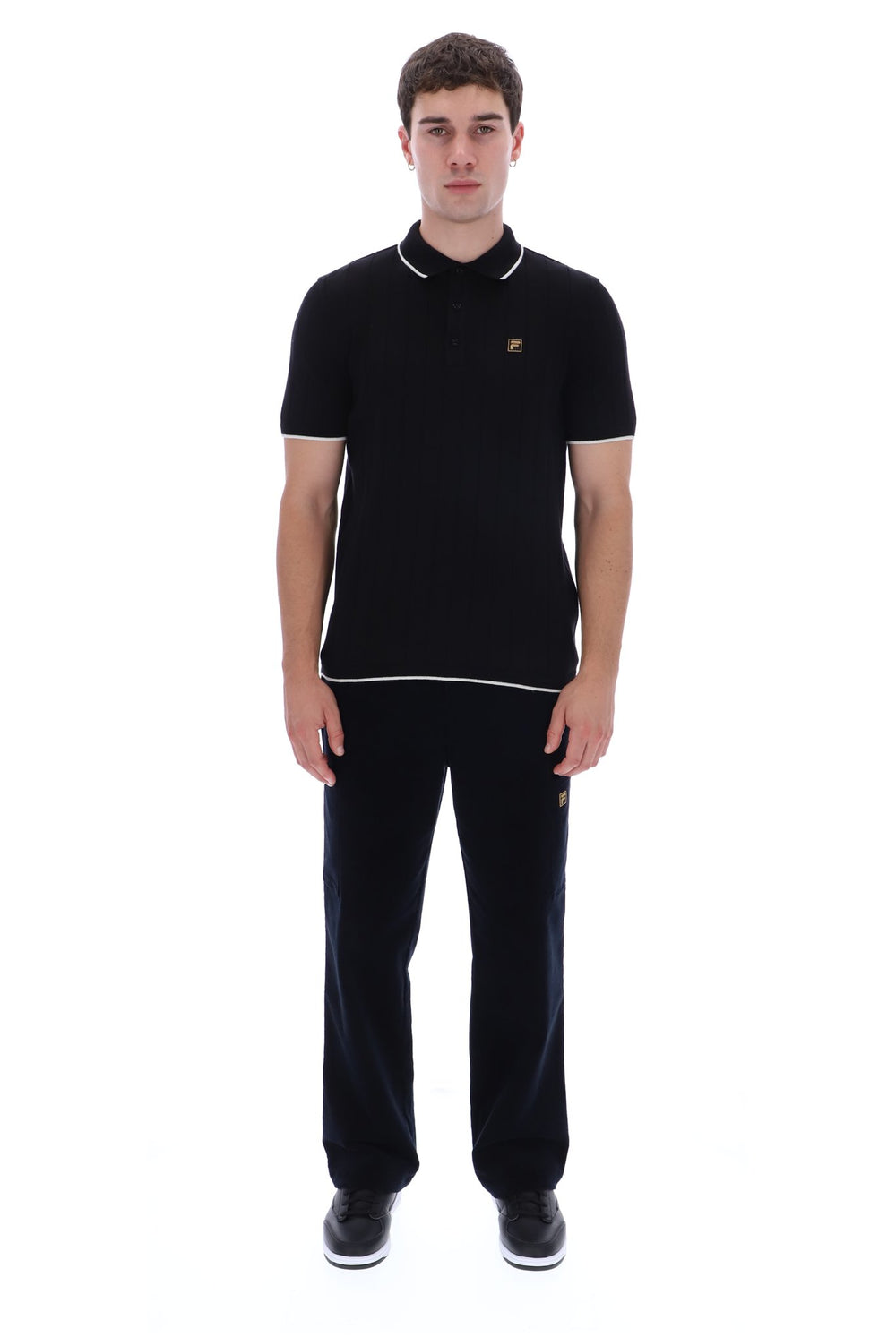 A man modeling a black short-sleeve polo shirt with white piping and a small gold embroidered logo on the chest, paired with black trousers and black sneakers, against a plain white background.