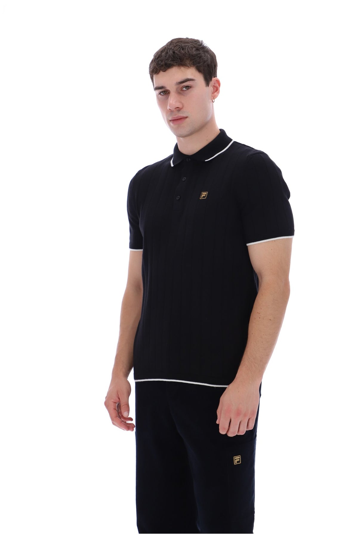 A man modeling a black short-sleeve polo shirt with white piping and a small gold embroidered logo on the chest, paired with matching black trousers, against a plain white background.