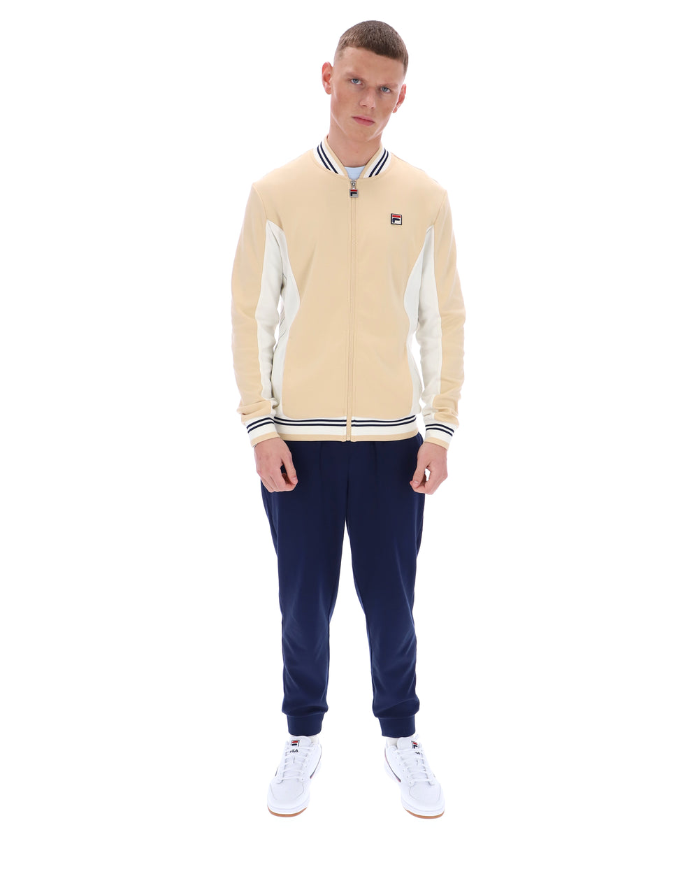 Man wearing a beige varsity jacket with white accents and navy pants on a white background