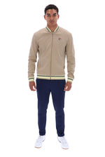 A man modeling a beige Fila zip-up bomber jacket with green and white striped ribbed collar, cuffs, and hem, paired with navy pants and white sneakers, on a solid white background.