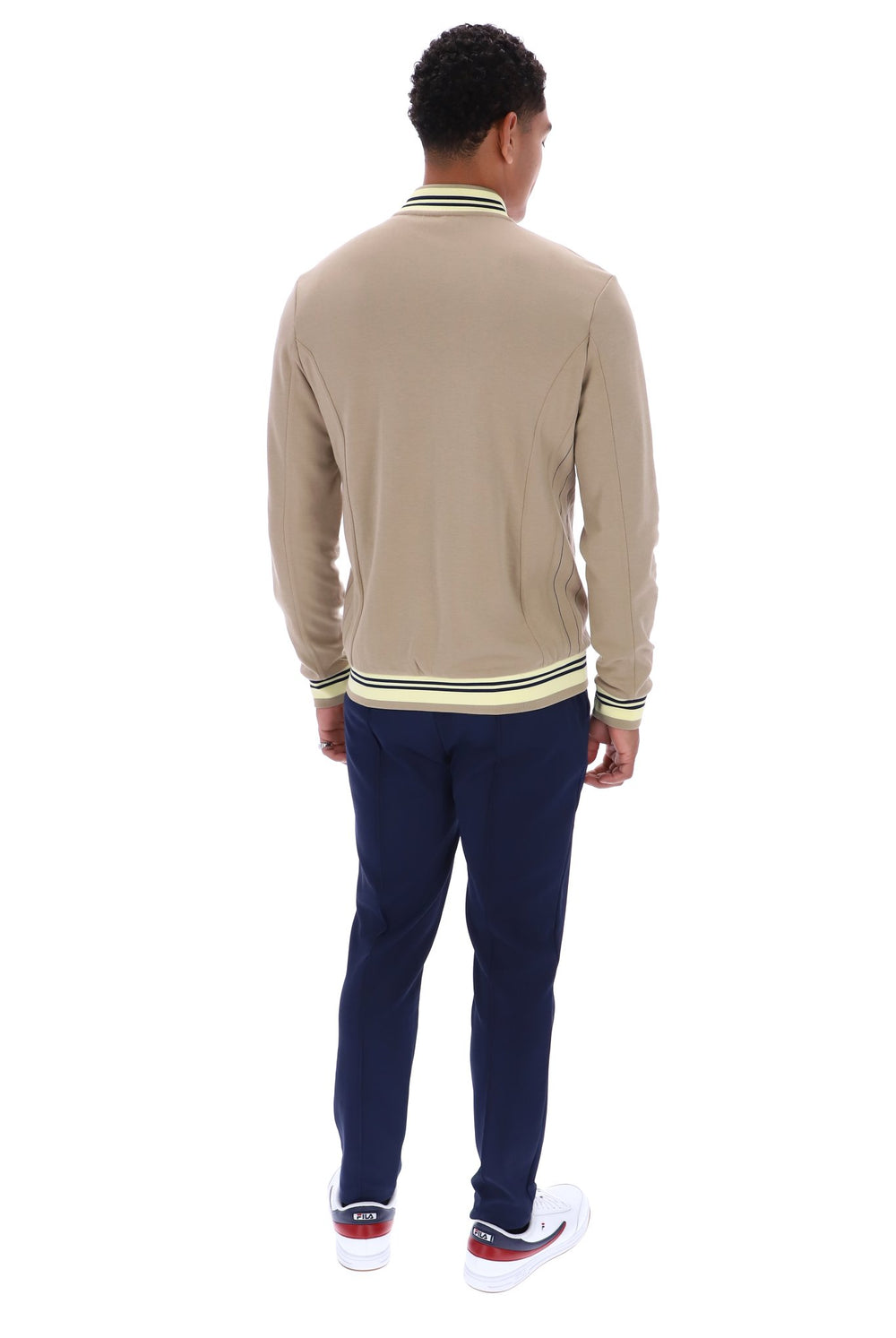A man viewed from behind wearing a beige long-sleeve knit sweater with green and white striped ribbed collar, cuffs, and hem, paired with navy pants and white sneakers with red accents, on a white background.