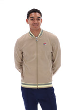 A man wearing a beige Fila zip-up bomber jacket with navy blue pants, photographed against a solid white background.