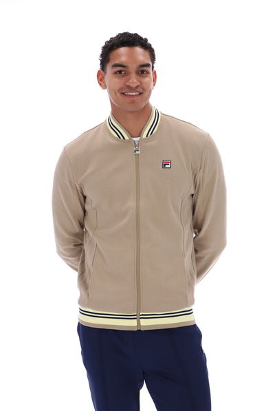 A man wearing a beige Fila zip-up bomber jacket with navy blue pants, photographed against a solid white background.