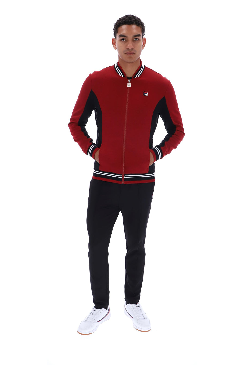 A man modeling a red and black zip-up bomber jacket with white and black striped ribbed collar, cuffs, and hem, paired with black pants and white sneakers against a plain white background.