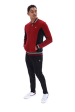 A man modeling a red and black Fila zip-up track jacket with striped ribbed collar, cuffs, and hem, paired with black sweatpants and white sneakers, on a solid white background.