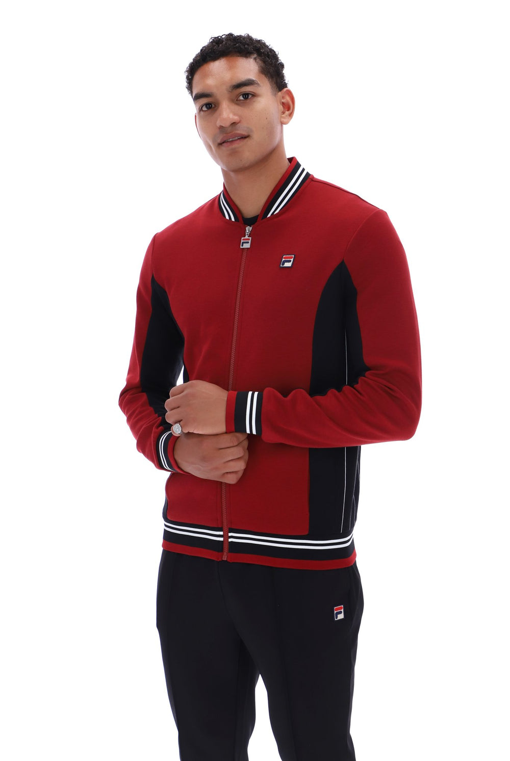 A man wearing a red and black Fila zip-up track jacket with white stripe details on the collar, cuffs, and hem, paired with black track pants, against a plain white background.