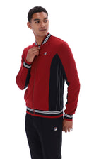 A man wearing a red and black Fila zip-up track jacket with white striped ribbed trim, paired with black track pants, against a plain white background.