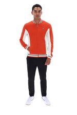 A man modeling a vibrant orange and white varsity-style zip-up jacket with striped ribbed collar, cuffs, and hem, paired with black pants and white sneakers against a plain white background.