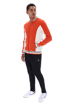 A man modeling a Fila zip-up jacket in orange and white, paired with black pants and white sneakers, against a solid white background.