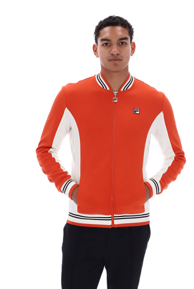 A man wearing a bright orange and white Fila zip-up varsity jacket with black and white striped ribbed collar, cuffs, and hem, paired with black pants, shot against a solid white background.