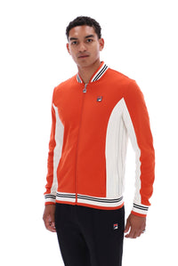 A man wearing a Fila orange and white colorblock zip-up track jacket with black track pants, photographed against a solid white background.
