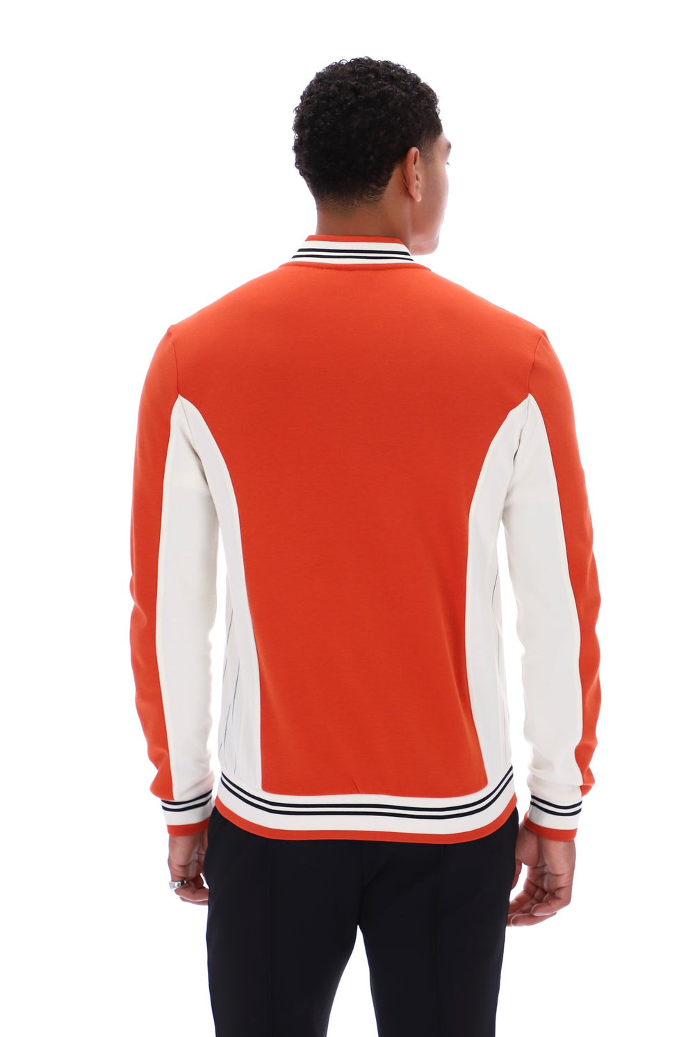 A man viewed from behind wearing a bright orange and white color-blocked knit sweater with striped ribbed collar, cuffs, and hem, paired with black pants against a plain white background.