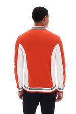 A man viewed from behind wearing a bright orange and white color-blocked knit sweater with striped ribbed collar, cuffs, and hem, paired with black pants against a plain white background.