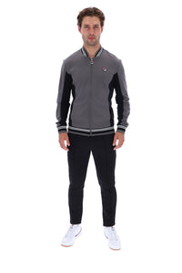 A man modeling a gray and black Fila zip-up track jacket with white and black striped ribbed collar, cuffs, and hem, paired with black pants and white sneakers, on a solid white background.