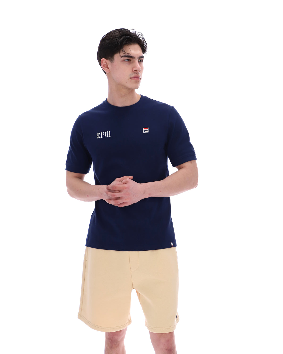 Man wearing a navy blue t-shirt with a FILA logo on the left side of the chest, and 'EST 1911' text on the right side of the chest. Paired with a beige shorts, against a white background