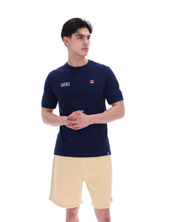 Man wearing a navy blue t-shirt with a FILA logo on the left side of the chest, and 'EST 1911' text on the right side of the chest. Paired with a beige shorts, against a white background