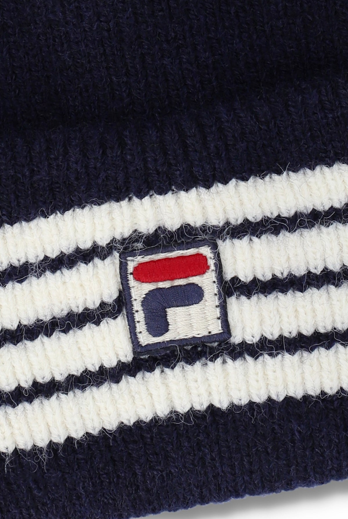 Close-up of a ribbed knit beanie in navy blue and white stripes, featuring a small embroidered Fila logo patch on the front.