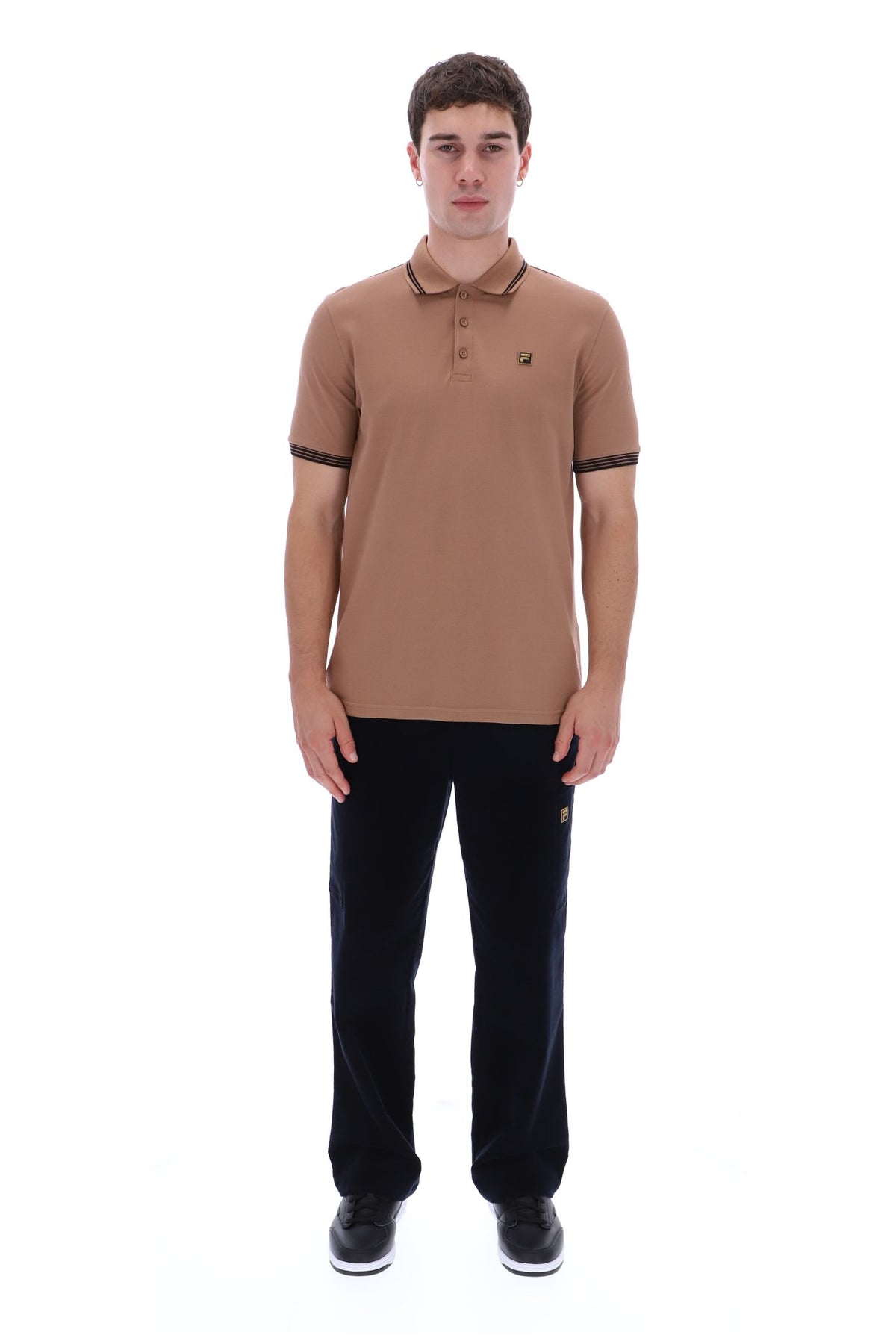A man modeling a tan short-sleeve polo shirt with dark trim on the collar and sleeve cuffs, paired with black trousers and black sneakers, against a plain white background.