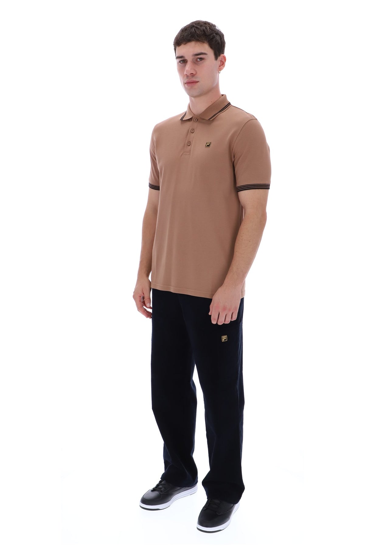 A man modeling a tan polo shirt with black sleeve trim and black trousers against a plain white background.