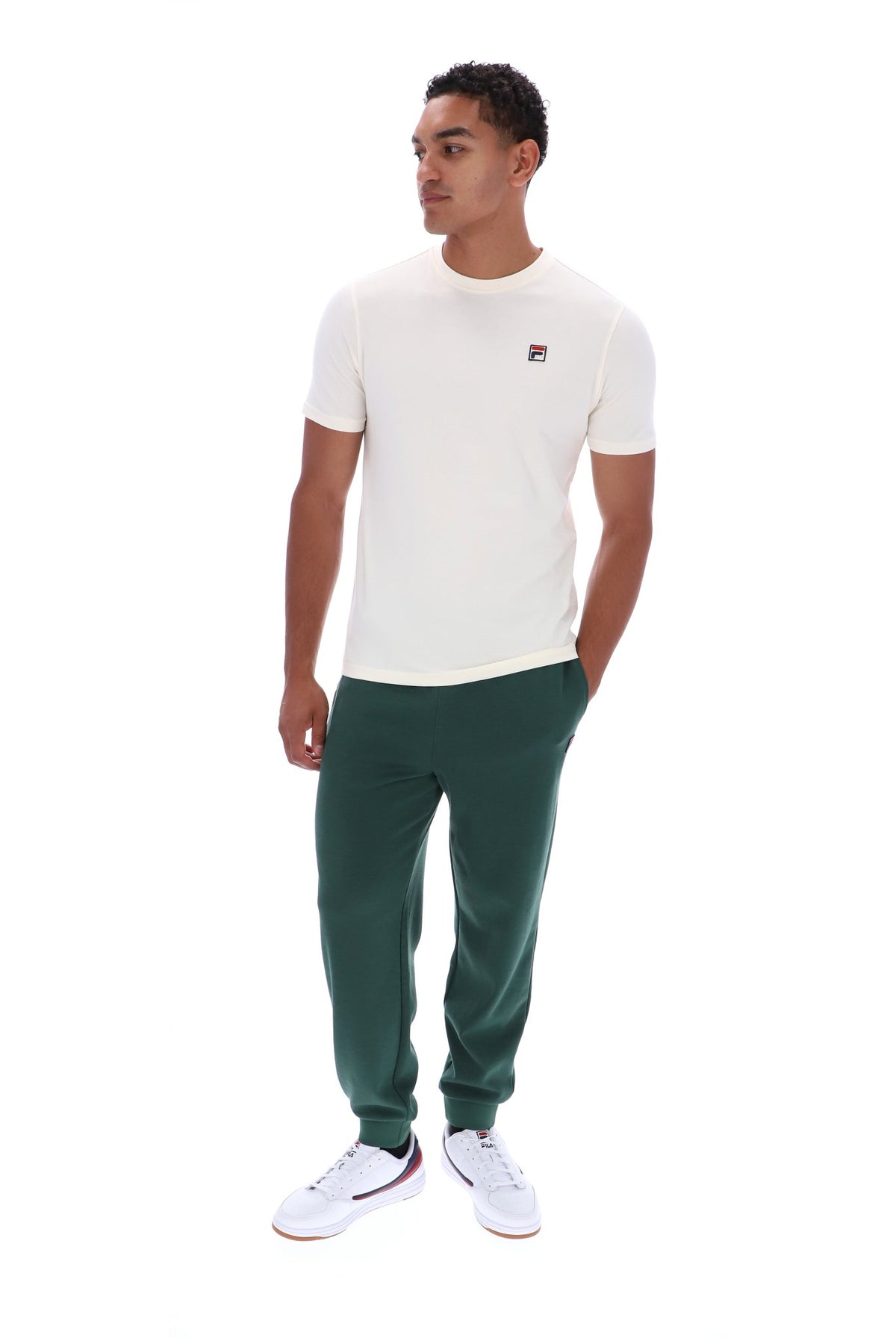 A man wearing a white short-sleeve crewneck t-shirt with a small red and blue Fila logo on the left chest, paired with dark green sweatpants and white sneakers, standing against a plain white background.