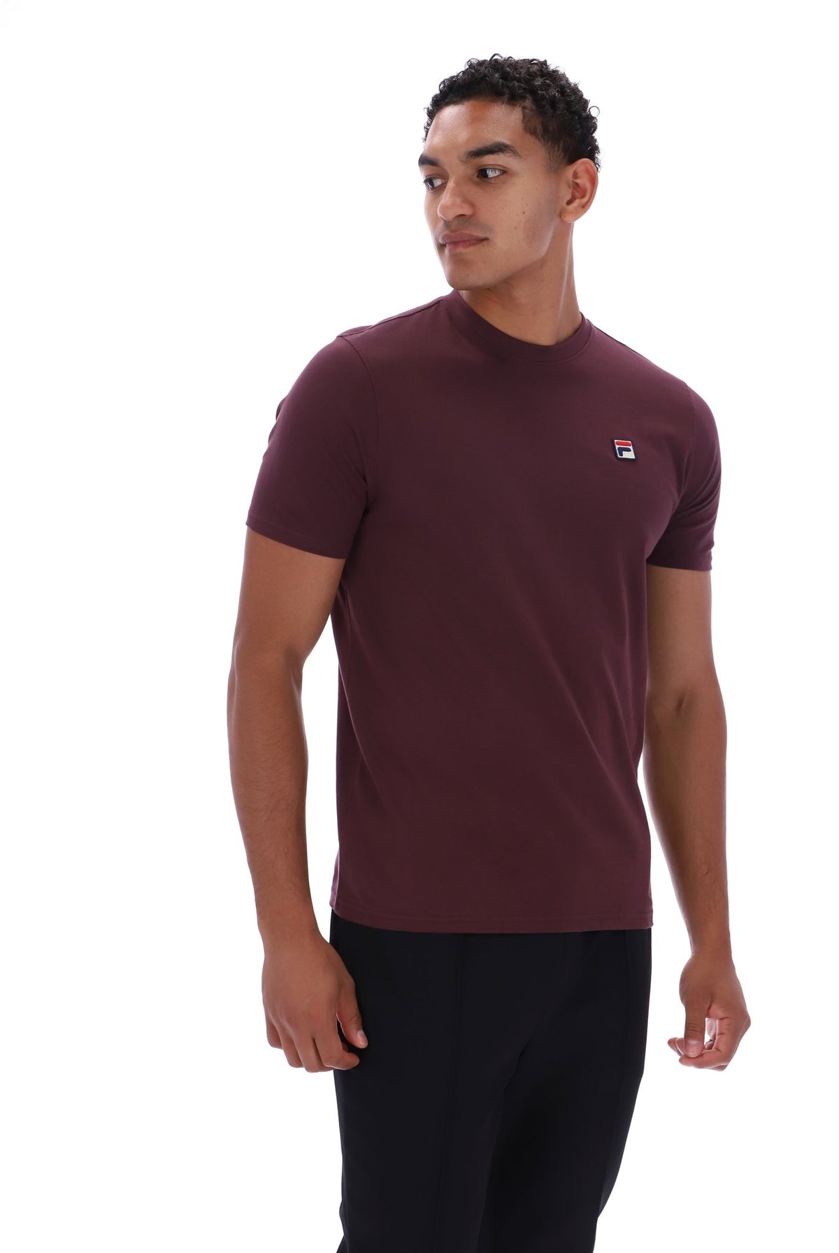 A man wearing a short-sleeve crewneck t-shirt in maroon with a small Fila logo on the left chest, paired with black pants, photographed against a solid white background.